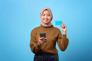 Feliz joven mujer asiática mostrando tarjeta de crédito y teléfono móvil en la mano sobre fondo azul.