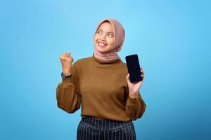 Feliz mujer asiática que muestra la pantalla en blanco del teléfono móvil y el éxito del gesto de la mano sobre fondo azul.