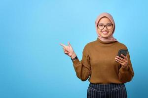 Feliz joven mujer asiática sosteniendo el teléfono móvil y señalar con el dedo para copiar el espacio sobre fondo azul.