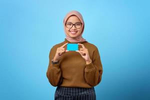 Retrato de joven alegre mujer asiática mostrando tarjeta de crédito sobre fondo azul.
