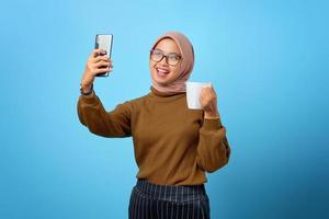 Alegre mujer asiática con teléfono móvil haciendo selfie y sosteniendo la taza sobre fondo azul.