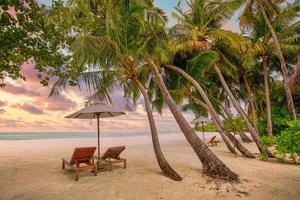 Romantic tropical sunset scenery, two sun beds, loungers, umbrella under palm tree. White sand, sea view with horizon, colorful twilight sky, calmness and relaxation. Inspirational beach resort hotel photo