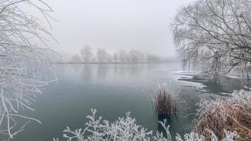 Winter forest and frozen lake sunset. Panoramic landscape with snowy trees, sun, beautiful frozen river with reflection in water. Cold winter landscape artistic foggy morning sunlight. Seasonal nature photo