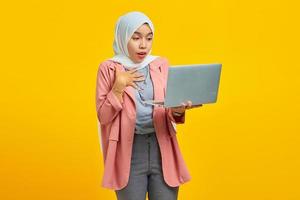 Portrait of a cheerful asian woman holding laptop and celebrating success isolated over yellow background photo