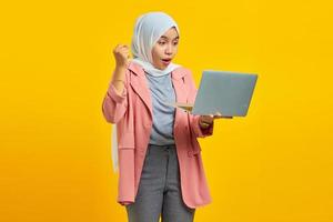 Portrait of a cheerful asian woman holding laptop and celebrating success isolated over yellow background