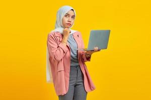 Portrait of beautiful young Asian woman holding laptop and pensive holding chin isolated over yellow background photo