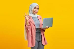 Portrait of cheerful young Asian woman holding laptop and showing ok gesture isolated over yellow background photo