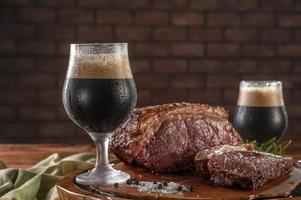 Two sweaty tulipa glasses of dark draft beer with grilled strip loin on wooden cutting board. Wooden table and bricks wall background. photo