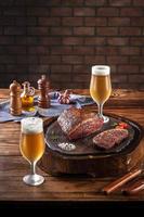 Grilled cap rump steak on wooden cutting board with two sweaty cold tulipa glasses of beer. Wooden table and bricks wall background  - brazilian picanha.