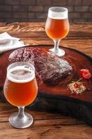 Grilled denver steak on wooden cutting board with two sweaty cold tulipa glasses of ale draft beer. Marble meat beef. photo