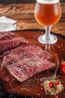 Grilled denver steak sliced on wooden cutting board with parrilla salt and sweaty glass of beer. Marble meat beef - Closeup.