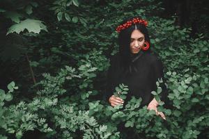 Beautiful girl in black dress and red decorations that holds a twig in her hand against the background of the forest. Space for your text message or promotional content photo