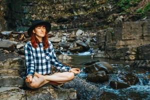 Woman with red hair in a hat and shirt meditating on rocks in a lotus position against a waterfall