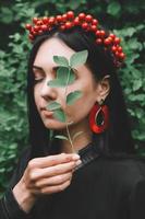 hermosa chica con cabello negro y accesorios rojos que sostiene una hoja en la mano cerca de su rostro contra el bosque foto