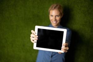 Mujer sonriente casual mostrando tableta digital con pantalla en blanco por pared verde foto