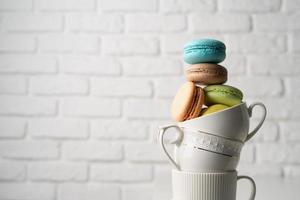 Stack of white coffee cups filled with macaroons on the edge of the table, white brick wall background photo