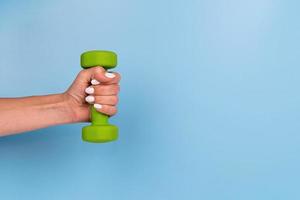 Female hand with white nails holding green dumbbell on blue background photo