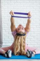 Athletic woman doing exercises using a resistance band on a fitness mat at home at white brick wall background photo