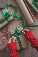 Manos de mujer sosteniendo un regalo de Navidad envuelto con papel artesanal, cinta verde y bastón de caramelo vista superior sobre mesa de madera foto