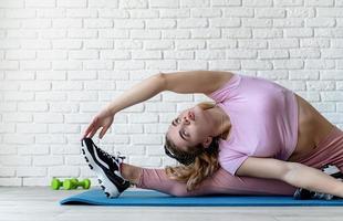 Mujer atlética que se extiende sobre una colchoneta de fitness en casa en el fondo de la pared de ladrillo blanco foto