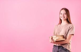 Happy teenager girl holding a pile of books isolated on pink background photo
