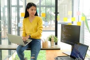 programadora asiática para camisa amarilla se sienta en el estante y coloca la computadora portátil en la pierna. Ella miró la pantalla de la computadora en la mesa y reflexionó. foto