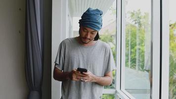 Asian man freelancer typing a message on his smartphone while standing beside the window in the office at home. video