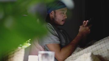 aziatische man die een mobiele telefoon gebruikt terwijl hij thuis op bed ligt.