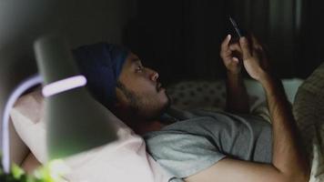 Apuesto hombre asiático usando un teléfono móvil mientras está acostado en la cama en su casa a altas horas de la noche. video