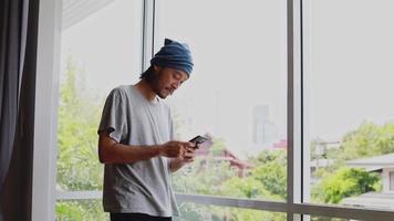 Apuesto hombre de negocios asiático escribiendo un mensaje en su teléfono inteligente mientras está de pie junto a la ventana en la sala de estar de su casa. video
