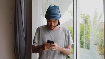 Freelancer de homem asiático sério digitando uma mensagem em seu smartphone em pé ao lado da janela em um escritório em casa.