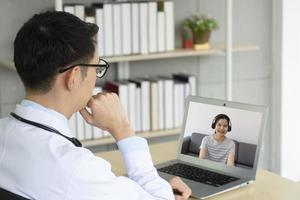 young asian doctor wearing headphone and using computer to give consult to the patient via video call in the hospital office. telemedicine and healthcare concept photo