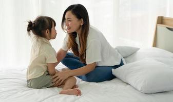asian mom and young kid with smiling face sitting together on bed in bedroom photo