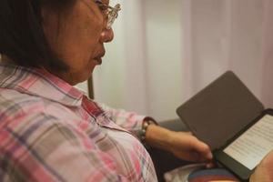 Senior reiteró a una mujer asiática leyendo un libro electrónico del e-reader en casa la noche antes de acostarse. ancianos usando el concepto de tecnología