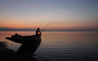 al lado del lago, pescador asiático sentado en un bote y usando caña de pescar para pescar al amanecer foto