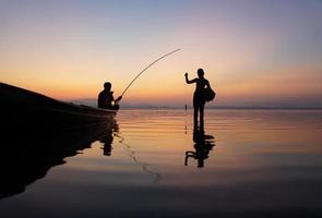 al lado del lago, un pescador asiático sentado en un bote mientras su hijo está de pie y usa una caña de pescar para pescar al amanecer