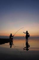 al lado del lago, un pescador asiático sentado en un bote mientras su hijo está de pie y usa una caña de pescar para pescar al amanecer foto