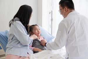 asian doctor using stethoscope to listen to heart of young girl who is lying in bed with mother in hospital pediatric ward. pediatrician and healthcare concept