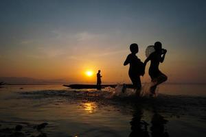 asian fisherman standing on boat and using fishing rod to catch fish while his son holding bamboo fish trap and running at lake side in morning photo