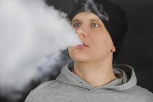 el hombre fuma un cigarrillo electrónico en el fondo oscuro, los hombres con capucha vapean y liberan una nube de vapor. chico con vaping sobre fondo negro foto