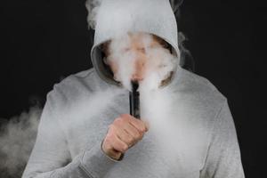 el hombre fuma un cigarrillo electrónico en el fondo oscuro, los hombres con capucha vapean y liberan una nube de vapor. chico con vaping sobre fondo negro foto