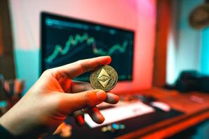 A man's hand holds an etherium coin at a computer table. photo