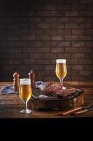 Bistec de grupa a la parrilla sobre tabla de cortar de madera con dos vasos de cerveza tulipa fría sudorosa. mesa de madera y fondo de pared de ladrillos - picanha brasileña. foto