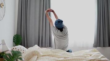 Asian man slowly waking up and stretches and gets up from the bed morning sun shines through the window at home. video