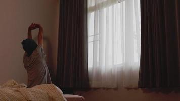 homem asiático acordando lentamente se estende e se levanta da cama para abrir as cortinas no início da manhã e olhando pela janela. video