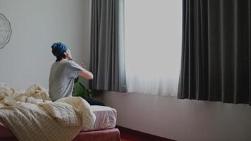 homem asiático acordando e se espreguiçando enquanto está sentado na cama, no quarto, o sol da manhã entra pela janela em casa.