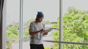 knappe aziatische man freelancer die foto op zijn tablet trekt terwijl hij naast het raam op kantoor thuis staat. video