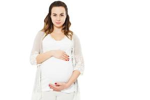 Bastante joven mujer embarazada de pie sobre fondo blanco y toca el vientre embarazado.