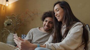 Young mixed race woman and young Middle Eastern man sitting on couch, woman holds phone, both waving goodbye, smiling to screen photo