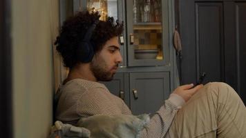 Young Middle Eastern man with headphone on his head, moves his head, sits, bent legs, holds and watches cell phone, sings a bit photo